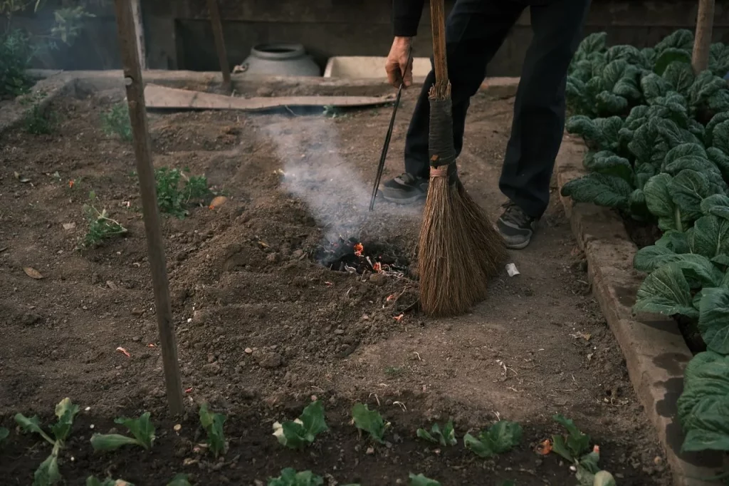 Jardinier brûlant des déchets végétaux dans une allée de potager, à côté d'un balai